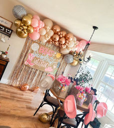 Playful cowgirl-chic bachelorette party setup in a sunlit dining room featuring pink, rose-gold and gold balloon garland, metallic fringe backdrop with a hand-lettered sign, pink cowboy hats and decorated gift bags on a table.