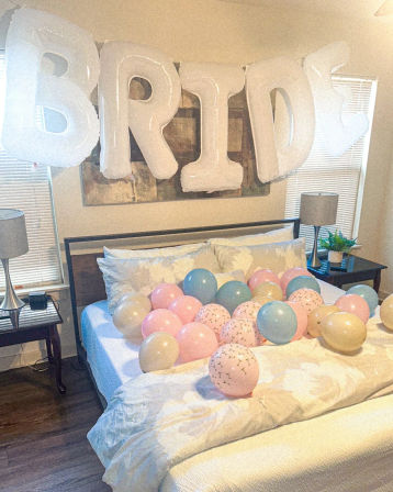 Bedroom styled for a bridal celebration with oversized white letter balloons spelling "BRIDE" above a bed covered in pastel pink, blue, and gold balloons on a white duvet, flanked by bedside tables with lamps and a small plant.