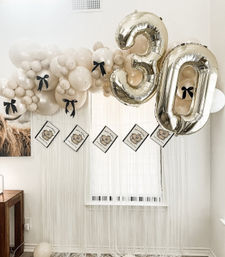 Neutral beige balloon garland accented with black bows and large gold foil "30" number balloons above a fringe window backdrop — elegant 30th birthday party setup in a home living room.