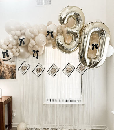 Neutral beige balloon garland accented with black bows and large gold foil "30" number balloons above a fringe window backdrop — elegant 30th birthday party setup in a home living room.