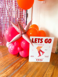 Indoor bachelorette party decor with hot pink and orange balloon cluster and metallic star spikes next to a 'LETS GO GIRLS' sign showing red cowgirl boots, pink sequin backdrop and hardwood floor — Fredericksburg, TX.