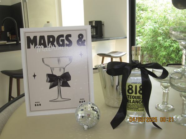 Kitchen bar tabletop with cocktail-themed poster, tequila bottle tied with a black ribbon, silver ice bucket, stacked glassware and a small disco-ball ornament — party-ready scene by a window with green bamboo outside.