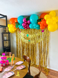 Fun tropical bridal-shower photo backdrop in a home dining room: gold fringe curtain with teal, orange, yellow and magenta balloon garland, faux leaves, table with novelty face fans and pink cups.