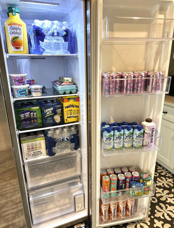 Open stainless-steel home refrigerator stocked with orange juice, bottled water, sparkling cans, coconut water cartons, ready-to-drink teas and packaged snacks on organized shelves — party-ready drinks in a modern kitchen.