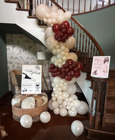Vineyard-themed entryway with a cream and burgundy balloon garland cascading down a wooden curved staircase, rustic ottoman, wine-inspired welcome signs, patterned wallpaper and warm hardwood floors.