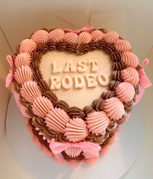 Heart-shaped pink and brown buttercream cake with piped rosettes, white "LAST RODEO" lettering and pink ribbon bows, playful western-themed celebration dessert.