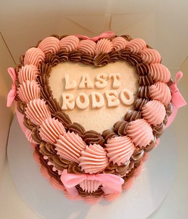Heart-shaped pink and brown buttercream cake with piped rosettes, white "LAST RODEO" lettering and pink ribbon bows, playful western-themed celebration dessert.