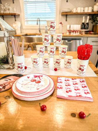 Cherry-themed party setup on a wooden kitchen island: pyramid of paper cups reading “cherry on top,” stacked plates, cherry-print napkins, red plastic utensils, striped paper straws and scattered decorative cherries.