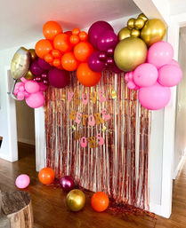 Indoor home party backdrop in a doorway: pink, orange, purple and gold balloon garland with a champagne glass foil balloon over a rose-gold metallic fringe curtain and letter banner, with matching balloons scattered on a hardwood floor.