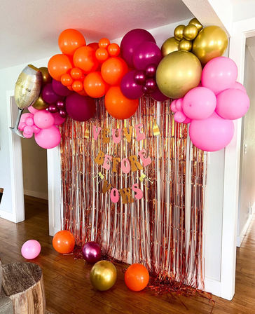 Indoor home party backdrop in a doorway: pink, orange, purple and gold balloon garland with a champagne glass foil balloon over a rose-gold metallic fringe curtain and letter banner, with matching balloons scattered on a hardwood floor.