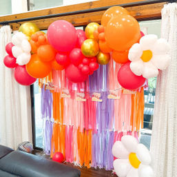 Bright indoor party backdrop featuring a pink, orange and gold balloon garland with oversized white daisy balloons and a multicolor tassel streamer curtain