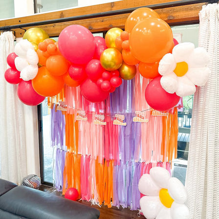 Bright indoor party backdrop featuring a pink, orange and gold balloon garland with oversized white daisy balloons and a multicolor tassel streamer curtain