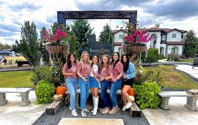 Six friends in matching shirts holding wine glasses at a vineyard estate entrance framed by hanging flower baskets, pumpkins and a white Mediterranean-style villa under a cloudy sky — outdoor wine tasting scene