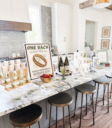 Bright modern farmhouse kitchen island with marble countertop set up for a bachelorette party—champagne bottles, flute tower, bowl of strawberries, disposable cups, decorative 'One Bach' sign with gold ring, and four metal bar stools.
