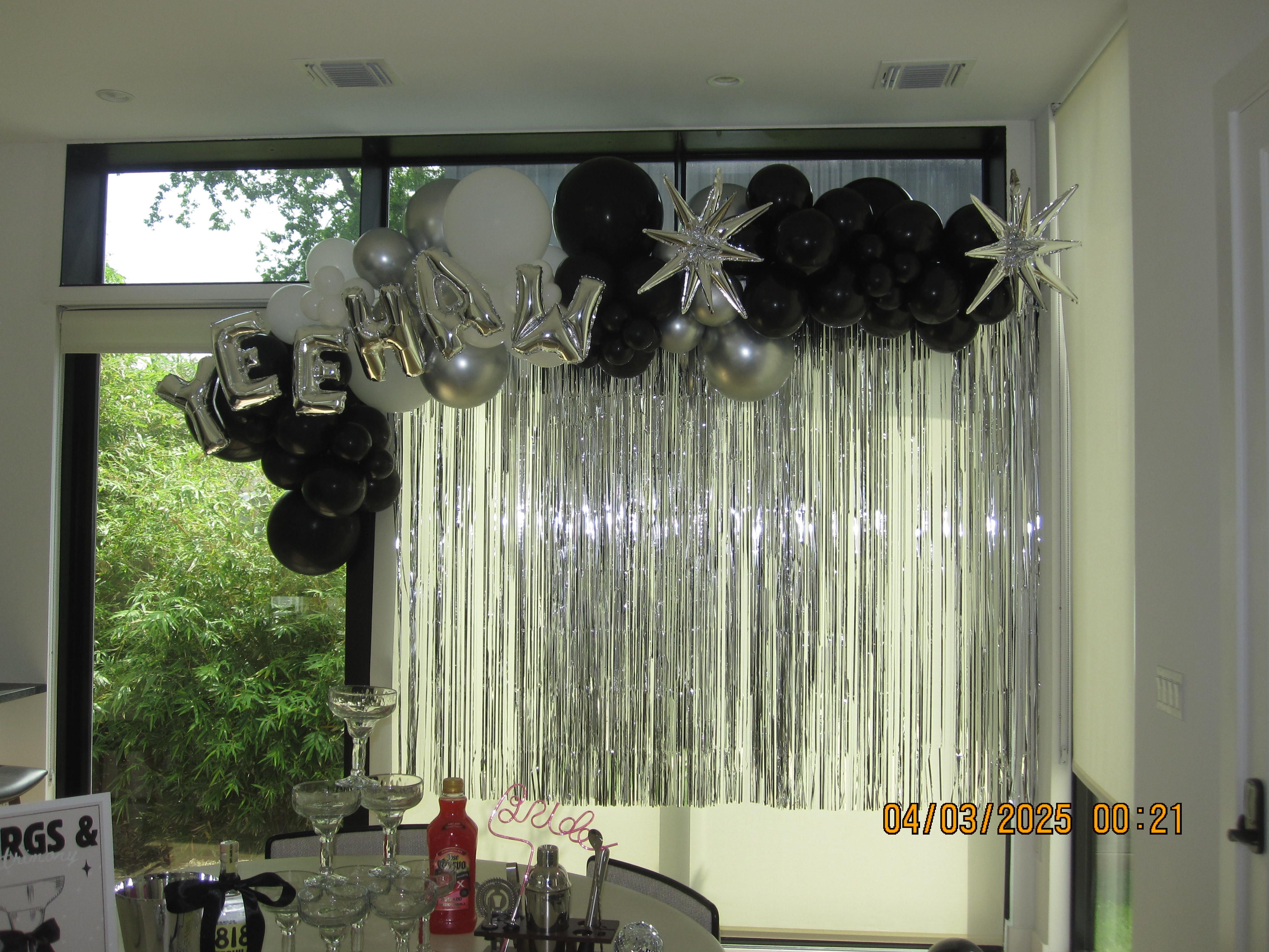 Sleek indoor party backdrop with black, white and silver balloon garland, star-shaped foil balloons and metallic silver fringe curtain above a table with a champagne glass tower and drink supplies