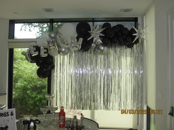 Sleek indoor party backdrop with black, white and silver balloon garland, star-shaped foil balloons and metallic silver fringe curtain above a table with a champagne glass tower and drink supplies