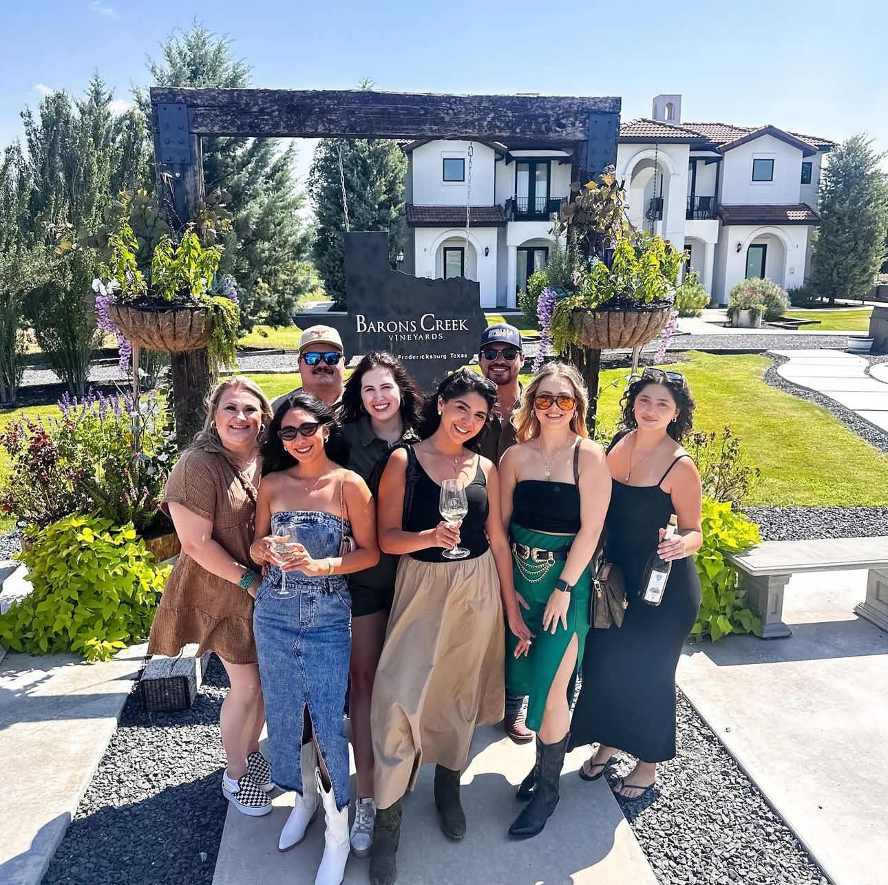 Eight friends smiling and holding wine glasses in front of a Mediterranean‑style tasting room at a sunny vineyard near Fredericksburg, Texas, with hanging flower baskets and manicured lawns.