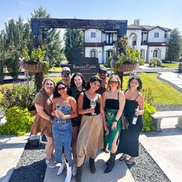 Eight friends smiling and holding wine glasses in front of a Mediterranean‑style tasting room at a sunny vineyard near Fredericksburg, Texas, with hanging flower baskets and manicured lawns.