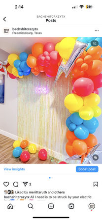 Bright rainbow balloon arch with silver star balloons, iridescent fringe backdrop and pink neon "love" sign over hardwood floor — colorful party setup in Fredericksburg, Texas.