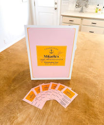 Pink-and-yellow champagne gift box with matching tasting cards fanned on a wooden kitchen island, bright white cabinets and door in the background.