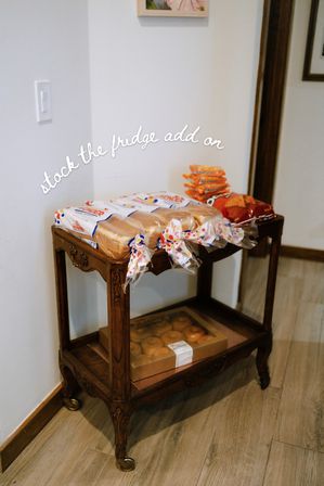 Vintage wooden rolling cart in a home entryway holding multiple loaves of sliced white bread, a box of donuts and stacked snack packs, with handwritten-style text saying 'stock the fridge add on'.