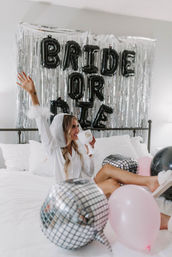 Bride-to-be lounging on a white bed in a veil and robe, holding a "Bride" goblet and waving in front of a silver fringe backdrop with black "BRIDE OR DIE" balloons, surrounded by disco-ball and pink balloons for a bachelorette party.
