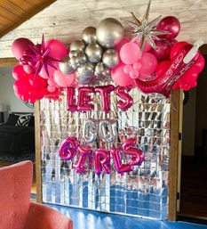 Indoor party photo wall with mirrored silver sequin backdrop, pink and metallic balloon garland, starburst accents, pink cowboy boot balloon and foil letters reading LETS GO GIRLS