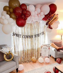 Cowboy-themed bachelorette setup in a living room with a gold fringe backdrop and slogan banner, pink, burgundy and cream balloon arch, gold ring and boot foil balloons, scattered balloons on hardwood floor and cushioned armchairs.