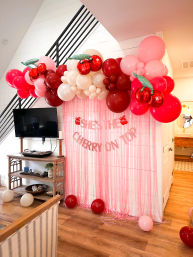 Cherry-themed indoor party backdrop in a living room with a pink fringe curtain, red-pink-cream balloon garland and cherry foil balloons, banner reading She's the cherry on top, and scattered balloons on a wood floor.