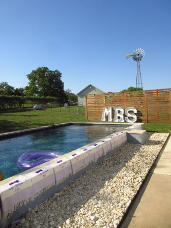 Backyard swimming pool with a purple inflatable ring, towels along the pool edge, white pebbles, large 'MRS' marquee letters by a wooden privacy fence and a tall farm windmill under a clear blue sky in a rural yard.