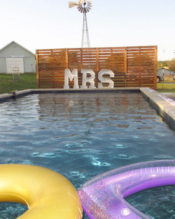 Backyard pool with gold and purple inflatable rings in the foreground, giant marquee letters spelling “MRS” against a wooden privacy screen and a windmill in the background.