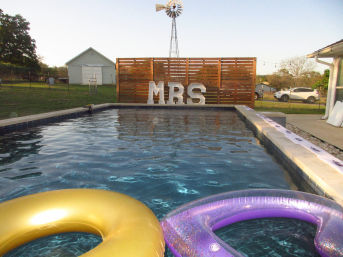 Rectangular backyard swimming pool with gold and purple inflatable rings in the foreground, a wooden slatted privacy screen displaying large 'MRS' marquee letters and a windmill in the background.