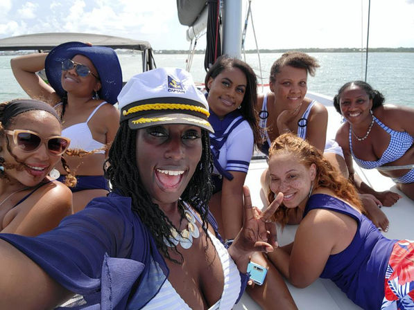 Seven friends in swimwear taking a joyful selfie on a sailboat deck, one wearing a captain's hat with blue sky and calm ocean in the background.
