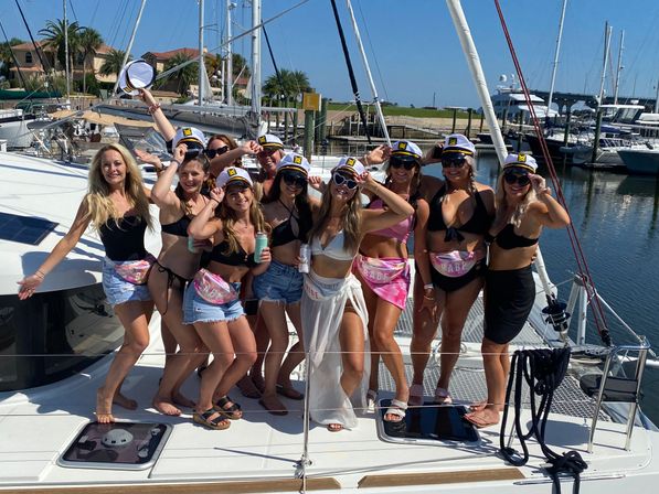 Group of women in swimsuits and captain hats posing on a sailboat deck at a sunny marina — bachelorette-style boat party with colorful fanny packs and drinks