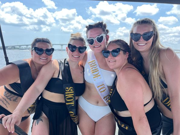 Five friends on a sunny boat bachelorette party in a blue bay, bride-to-be wearing a white sash and heart sunglasses, group in black swimsuits smiling with a coastal bridge and fluffy clouds in the background.