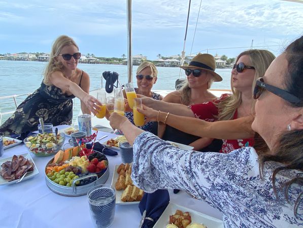 Group of women toasting mimosas over a sunny sailboat brunch with a fruit platter and coastal waterfront homes in the background.