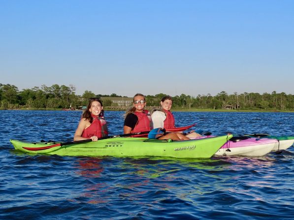 Three smiling people in red life jackets kayaking in bright green and pink kayaks on a sunlit blue lake with a tree-lined shoreline and clear sky — outdoor summer paddling scene.