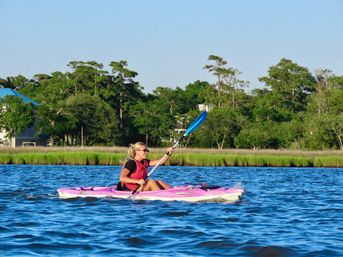 Smiling kayaker in a pink kayak paddling with a blue paddle across bright blue estuary waters past grassy salt marsh and pine-lined shoreline under a clear sky.