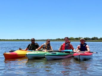 Four kayakers in colorful kayaks paddling on calm coastal marsh water under a bright blue summer sky, wearing life jackets and smiling