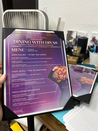 Vibrant purple laminated event menu titled "Dining with Divas" showing appetizers, main acts and pizza performances, with a colorful drag performer illustration; menu held in a black corner folder resting on a cluttered desk with a fan, pens and papers nearby.