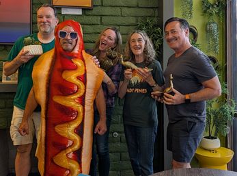 Playful indoor group photo of five adults—one in a hot dog costume—smiling and holding brass animal figurines and a potted succulent against a green brick, plant-filled wall.