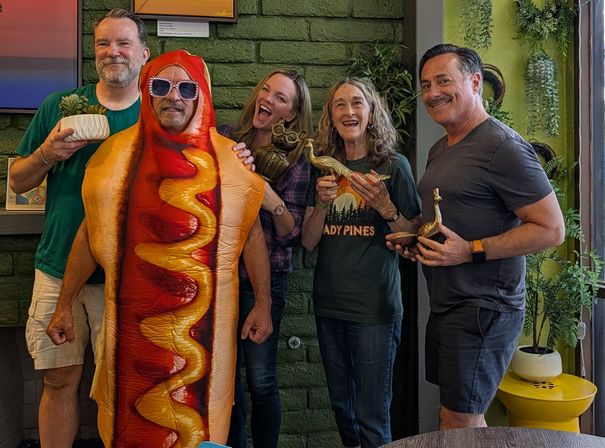 Playful indoor group photo of five adults—one in a hot dog costume—smiling and holding brass animal figurines and a potted succulent against a green brick, plant-filled wall.
