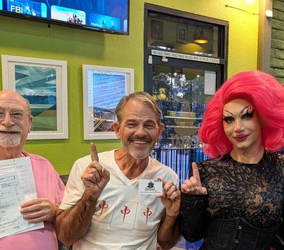 Four smiling adults pose inside a colorful cafe with green walls and framed art — a drag performer in a bright pink wig holds a prize card while two others show a trivia night answer sheet and all point up with one finger.