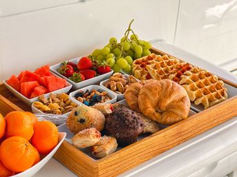 Colorful breakfast tray with golden waffles, flaky croissant, chocolate and blueberry muffins, fresh green grapes, strawberries, watermelon cubes, tangerines, mixed nuts and cereal bits on a wooden serving tray on a white countertop.