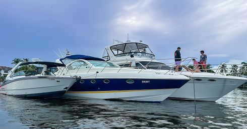 Three luxury motor yachts moored side-by-side at a palm-lined marina, friends socializing on the bow under a cloudy blue sky with calm water reflections