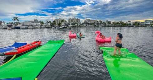 People enjoying bright green floating mats and pink flamingo inflatables on calm water at a palm-lined waterfront marina with boats and condos under a blue cloudy sky.