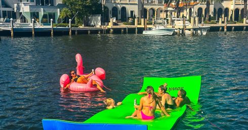 Group of people lounging on a bright green inflatable water mat and a pink flamingo float in a sunny waterfront canal lined with luxury homes and docked boats.