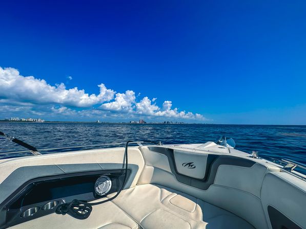 View from a boat bow with inviting white seats over calm blue ocean toward a distant coastal skyline under a bright blue sky dotted with fluffy clouds.
