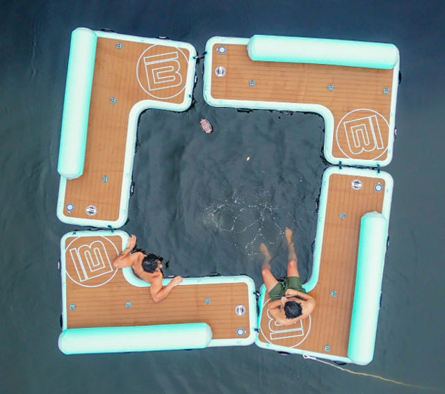 Aerial view of a square inflatable floating dock with teak-style decks and mint-blue bumpers on dark lake water, two people lounging on the edges with legs in the water and one person swimming in the center — summer lake recreation.