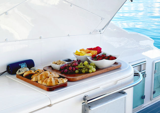 Yacht breakfast spread on a white boat deck with calm blue sea in the background — wooden boards with croissants and pastries, bowls of strawberries, melon and watermelon, clusters of red and green grapes, and a small dish of nuts and berries.